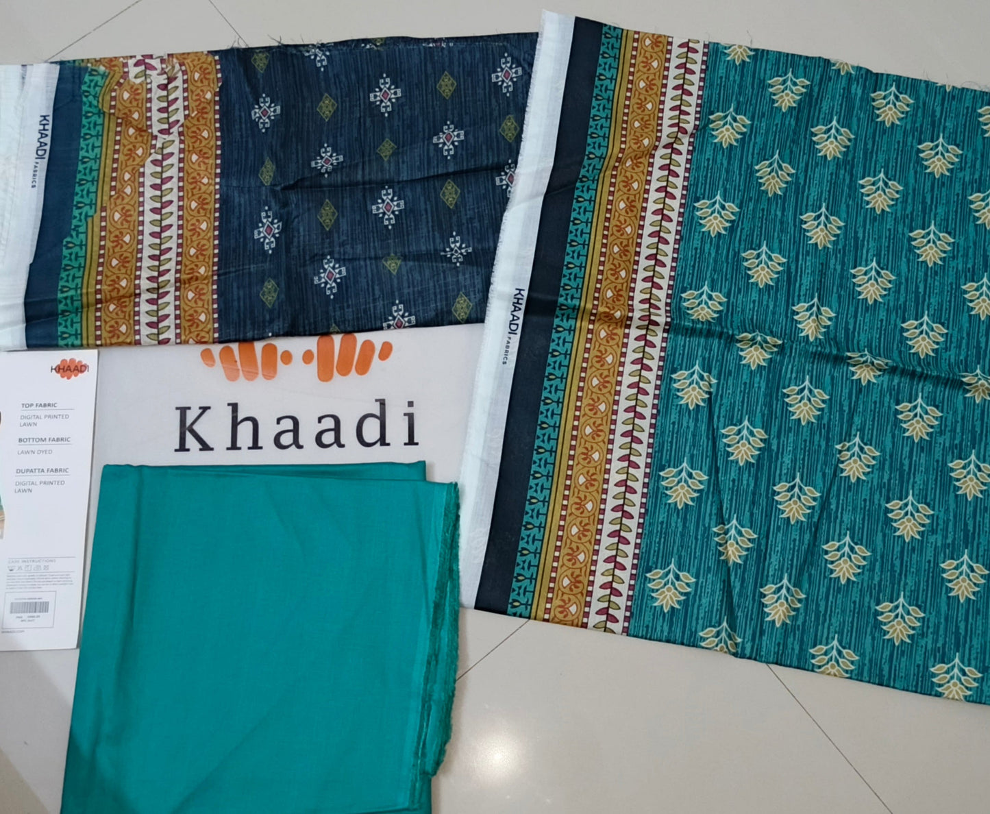 Khaadi fabric samples on a tiled floor with promotional material.